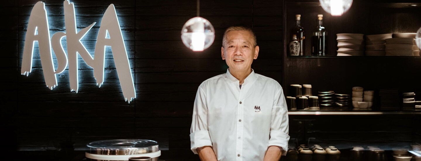 Ein Küchenchef steht hinter einer Sushibar in einem Restaurant mit einem Wandlogo Aska.