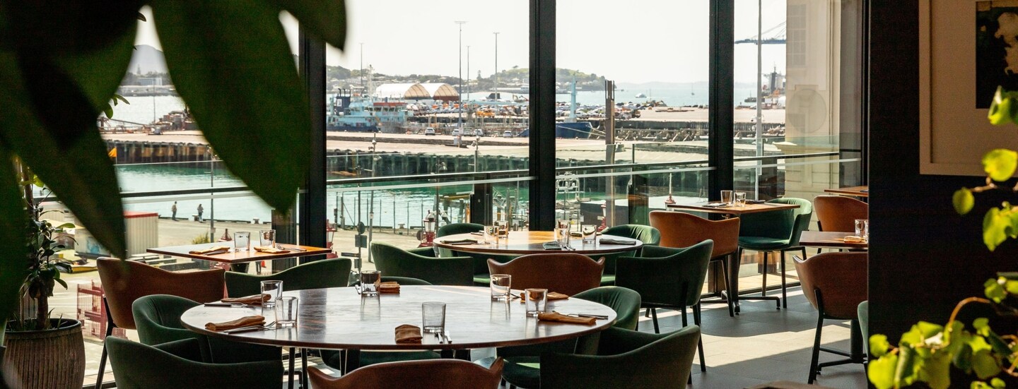 Runde Esstische in einem Restaurant mit Blick durch Panoramafenster auf ein Hafengebiet.