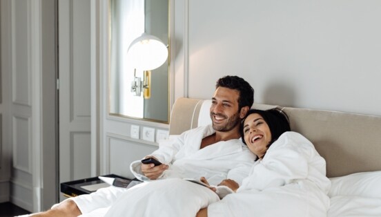 Ein fröhliches Paar in weißen Bademänteln entspannt im Bett einer edlen Hotelsuite