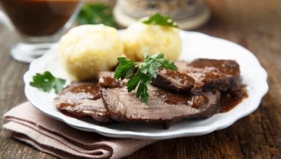 Sauerbraten mit brauner Soße an Klößen, angerichtet auf einem weißen Teller auf einem Holztisch.