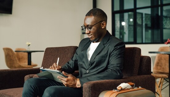 Ein Geschäftsmann arbeitet an einem Tablet auf einer Couch in einer Lounge, neben ihm steht ein Koffer.