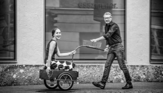 Ein Mann zieht eine lächelnde Frau in einem Fahrradanhänger auf einem Gehweg vor einer Fensterfront.