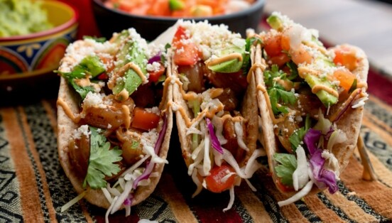 Drei aufgereihte Tacos, die mit Shrimps, Avocado, Koriander und mehr gefüllt und mit Soße angerichtet sind.
