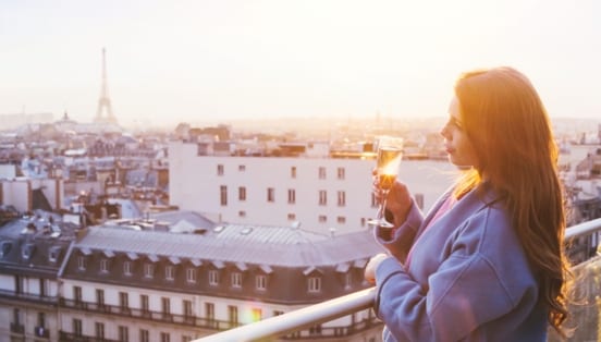 Eine Frau, die bei Sonnenuntergang in Paris auf einem Balkon steht und ein Glas Sekt in der Hand hält.