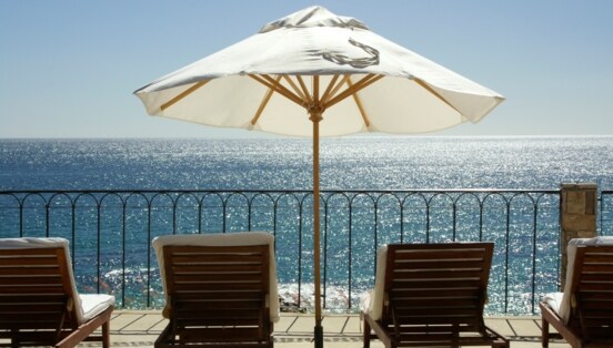 Terrasse und Geländer mit Blick aufs Meer, auf der mehrere Liegestühle und ein weißer Sonnenschirm stehen.