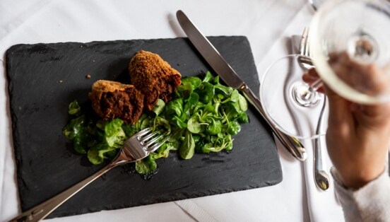 Aufsicht eines panierten Fleischgerichtes an Feldsalat auf einer Schieferplatte auf einem edel gedeckten Tisch, im Vordergrund eine Hand mit einem Glas Weißwein.