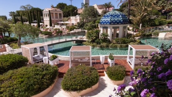 Ein lang gestreckter, mit Palmen, Blumen und Daybeds gesäumter Pool, über den zwei Brücken führen.