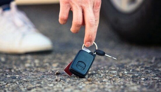 Nahaufnahme einer Hand, die nach einem Autoschlüssel am Asphaltboden greift, dahinter ein Autoreifen und Sneaker in Unschärfe.