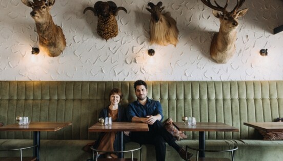 Anna und Nick sitzen am Tisch auf einem grünen Liedersofa. Oben an der Wand hängen Hirschköpfe.