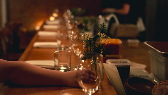 Gedeckter Holztresen bei schummrigen Licht, auf den eine Frauenhand eine kleine Blumenvase mit Kamille stellt.