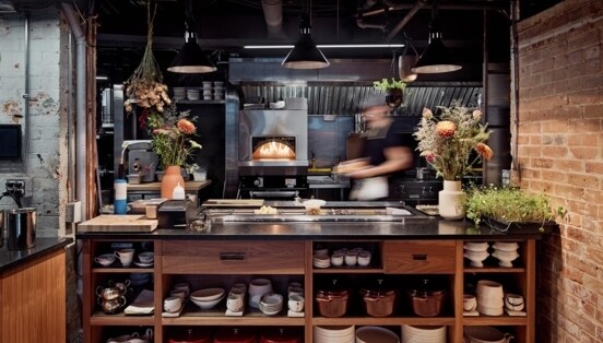 Blick in die Küche, wo im Ofen ein Feuer brennt und neben der unverputzten Backsteinwand eine Anrichte mit Blumensträußen und frischen Kräutern steht.