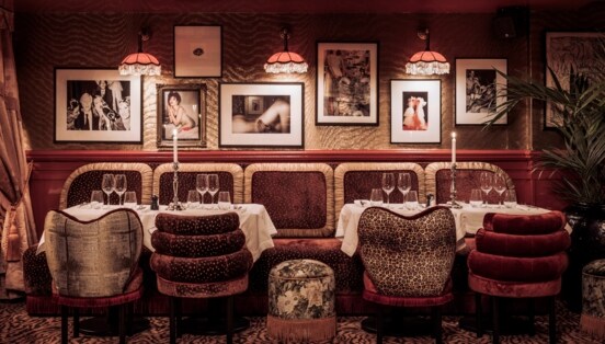 Gedeckte Tische in einem in dunklen Farben gehaltenem Interieur mit Sesseln und Teppich mit Animal-Muster sowie Fotografien an der Wand und Fransenlampen an der Decke.