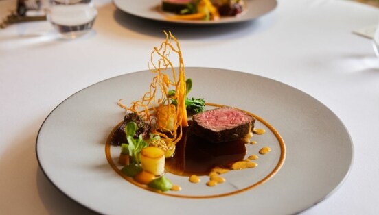 Kunstvoll arrangiertes Gericht mit einem Stück Fleisch auf einem großen weißen Teller auf einem weißen Tisch.