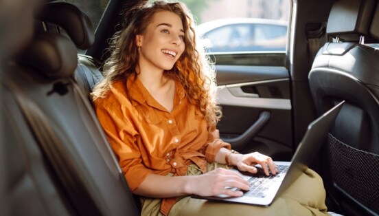 Eine lachende Frau in orangefarbener Bluse sitzt auf dem Rücksitz eines Autos mit einem aufgeklappten Laptop auf ihrem Schoß.