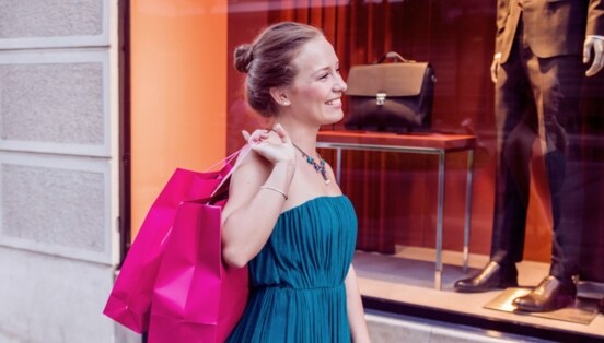 Eine lächelnde Frau im Kleid steht vor dem Schaufenster einer Modeboutique und hält zwei pinke Einkaufstüten über ihrer Schulter.