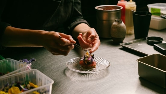 Zwei Hände garnieren eine edle Vorspeise mit Blüten auf einer Küchentheke.