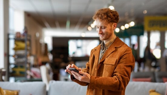 Ein junger Mann steht in einem Möbelgeschäft und schaut lächelnd auf das Smartphone in seiner Hand.