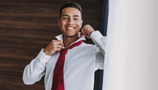 Ein junger Mann im weißen Hemd mit roter Krawatte richtet sich lächelnd den Kragen vor einem Spiegel.