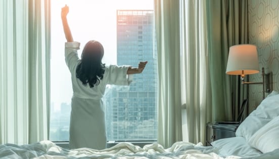 Rückansicht einer sich räkelnden Person im weißen Bademantel am Fenster eines Schlafzimmers in einer Großstadt, im Vordergrund ein Bett.