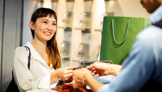 Eine lächelnde Frau bezahlt mit einer Kreditkarte bei einer Person in einem Geschäft, neben ihr eine grüne Einkaufstasche auf der Verkaufstheke.