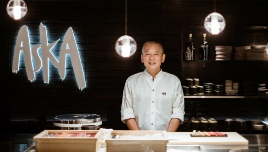 Ein Küchenchef steht hinter einer Sushibar in einem Restaurant mit einem Wandlogo Aska.