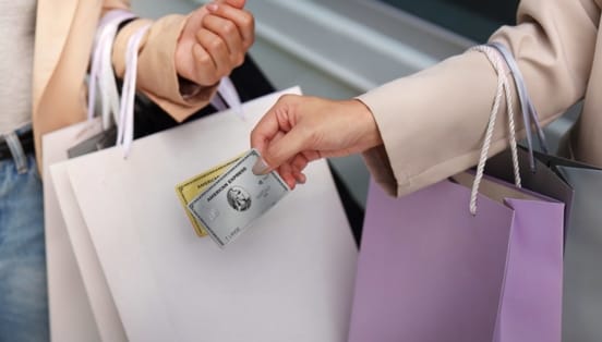 Nahaufnahme von zwei Armen mit Einkaufstüten, eine Hand hält eine goldene und eine silberne Kreditkarte von American Express.