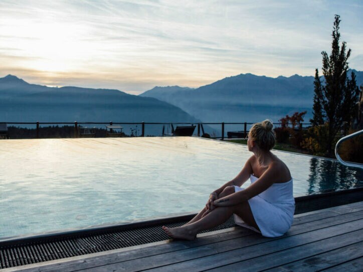 Eine Frau mit umwickelten Handtuch sitzt an einem Outdoor-Pool mit Blick auf die Berge in der Dämmerung
