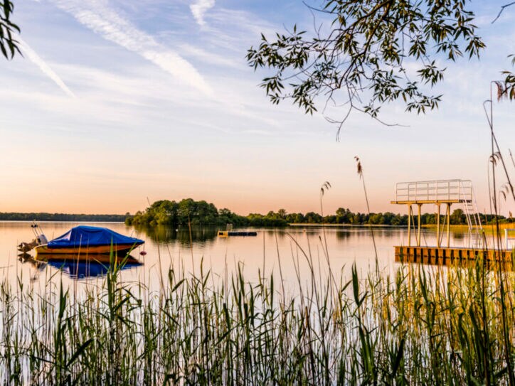 Ein See mit einem Boot und einem Sprungturm in der Abendsonne, im Vordergrund Schilf