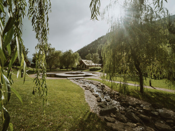 Ein mit Steinen gesäumter Bachlauf, der zwischen Bäumen in die Richtung eines Hauses im Hintergrund fließt.