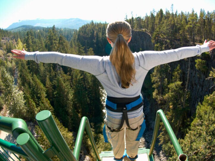 Eine Frau steht mit ausgebreiteten Armen gesichert an einem Bungee-Jumping-Gerüst und ist kurz davor runterzuspringen