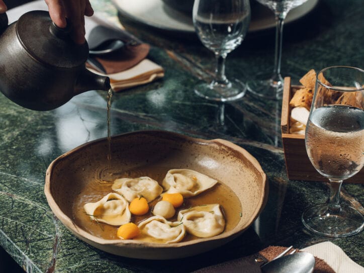 An einem stilvoll gedeckten Restauranttisch läuft Brühe aus einer Kanne in einen Teller mit gefüllter Pasta
