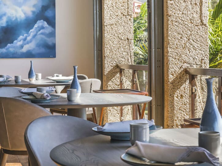 Innenraum eines luftigen, hellen Restaurants mit gedeckten Tischen an einer Fensterfront mit Blick auf Palmen