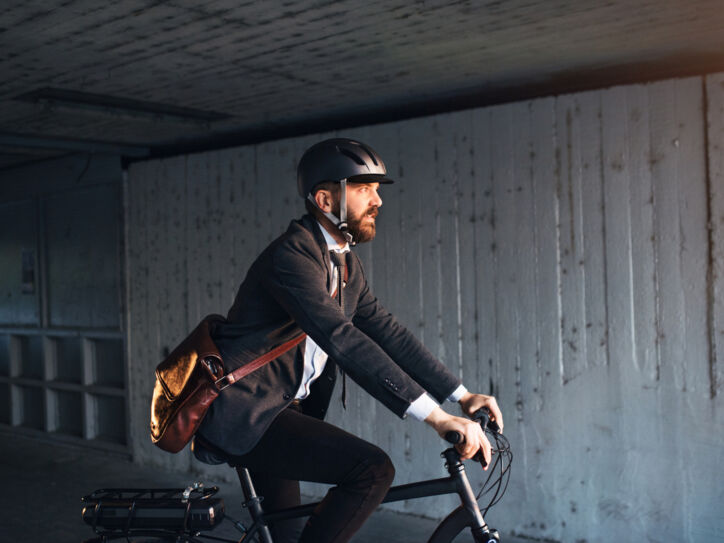 Seitenansicht einer Person im Anzug, mit Umhängetasche und Helm, die auf einem Fahrrad fährt