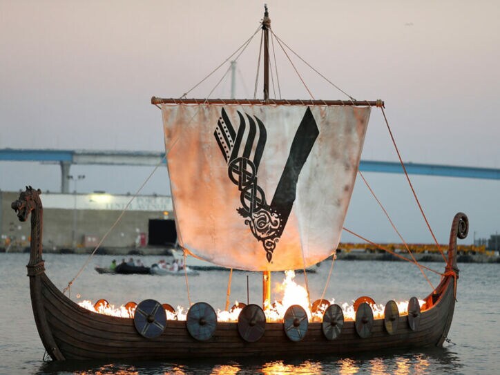 Aufnahme eines brennenden Wikingerschiffs auf dem Meer mit einem Segel, das das Logo der „Vikings“-Serie zeigt