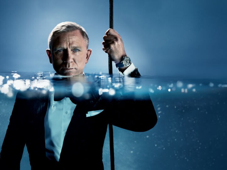 Porträt von Daniel Craig im schwarzen Smoking im Wasser, das ihm bis zum Hals steht; an seiner hochgehaltenen Hand trägt er eine Taucheruhr der Marke OMEGA