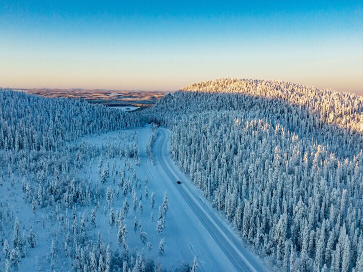 Luftaufnahme einer weiten Winterlandschaft mit schneebedeckten Wäldern, durch die ein einzelnes Auto über eine Straße durch den Schnee in der Abenddämmerung fährt