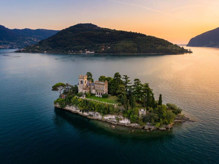 Luftaufnahme einer kleinen Felsinsel mit Burg und Bäumen in einem großen See bei Sonnenuntergang