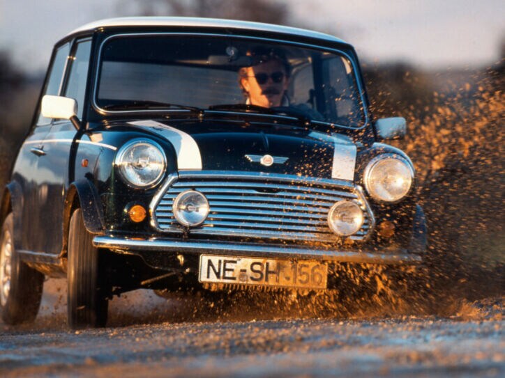 Ein Mann fährt in einem schwarzen MINI Cooper 1.3 mit Rallystreifen durch eine Schlammpfütze