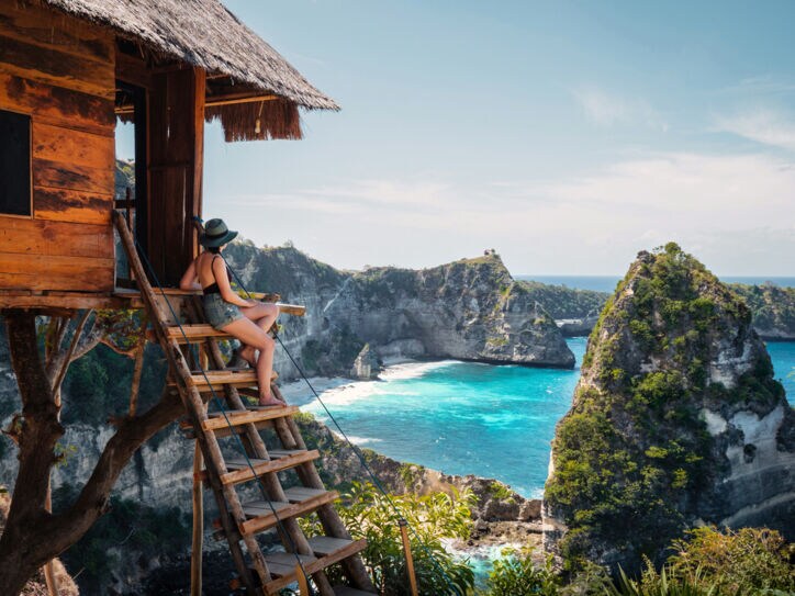 Rückansicht einer Frau mit Sonnenhut, die vom Eingang eines Baumhauses auf einem Felsen hinunter auf eine idyllische Bucht mit Sandstrand blickt