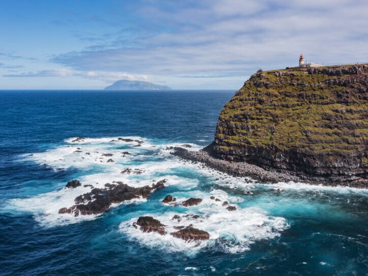 Luftaufnahme einer Steilklippe mit Leuchtturm und der Küste vorgelagerte Felsen im Meer, im Hintergrund eine Insel