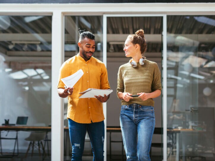 Zwei lässig gekleidete Personen stehen mit Arbeitsmaterialen vor einem offenen Büro mit einem langen Tisch.