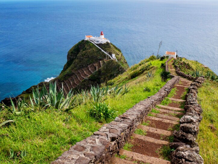 Blick von einem grasbewachsenen Hügel auf einen zweiten Hügel mit Leuchtturm auf einer Insel vor Meereskulisse.