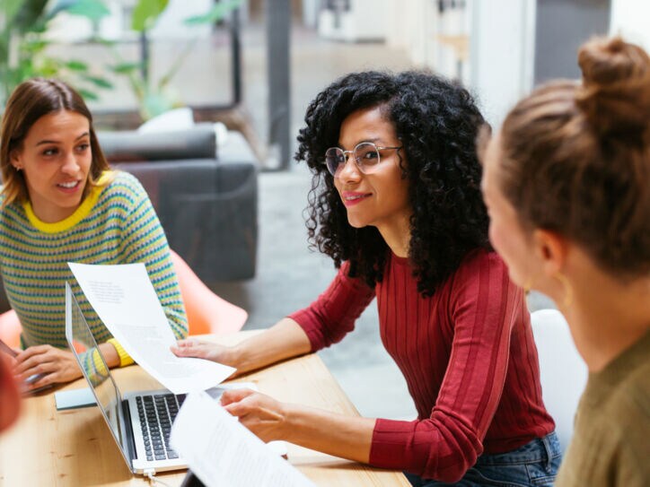 Drei junge Frauen sitzen mit Arbeitsunterlagen zusammen an einem Holztisch in einem Co-Working-Space.