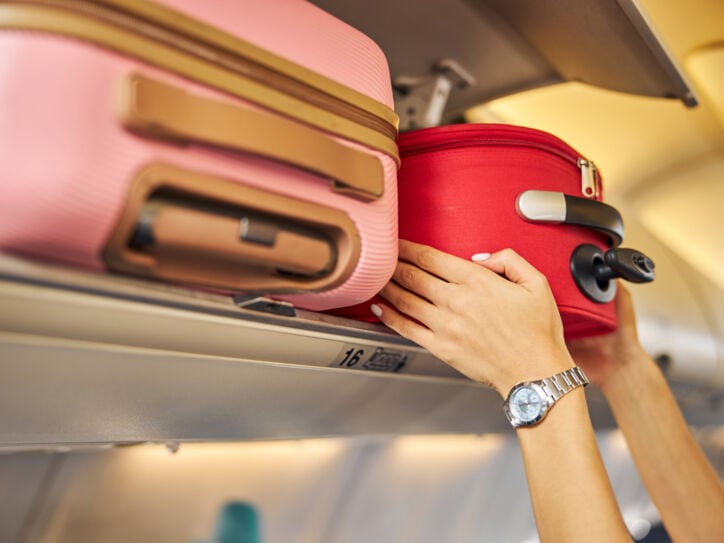 Zwei Hände greifen einen roten Koffer in einem Overhead-Bin eines Flugzeugs, im Vordergrund ein Koffer in Rosa.
