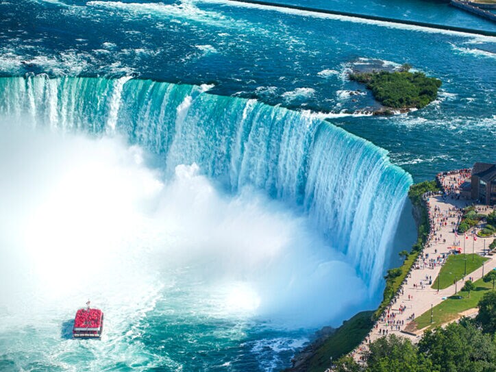Luftaufnahme eines runden Wasserbeckens mit Boot vor einem Wasserfall, am Ufer eine Aussichtsplattform mit Personen.
