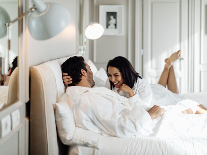 Ein Paar in weißen Bademänteln liegt lachend in einem weißen Bett in einem edlen, hellen Zimmer.