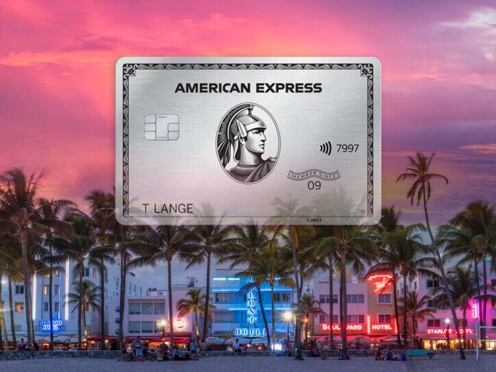 Freisteller einer Platinum Card von American Express vor der erleuchteten Oceanfront von Miami Beach hinter Palmen bei Sonnenuntergang.