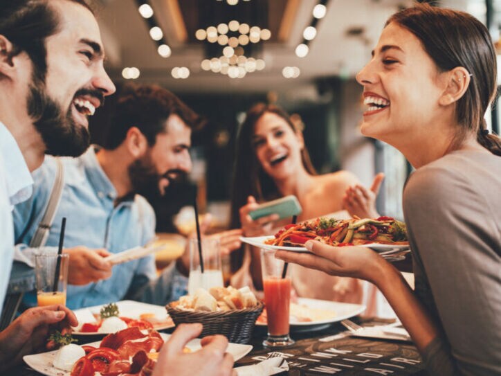 Eine Gruppe fröhlicher Personen in einem Restaurant. Eine lachende Frau hält einen Teller in der Hand.
