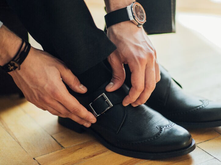 Männerhände schließen den seitlichen Schnallriemen eines schwarzen Monkstrap-Schuhs mit Lochmusternaht auf der Zehenkappe.