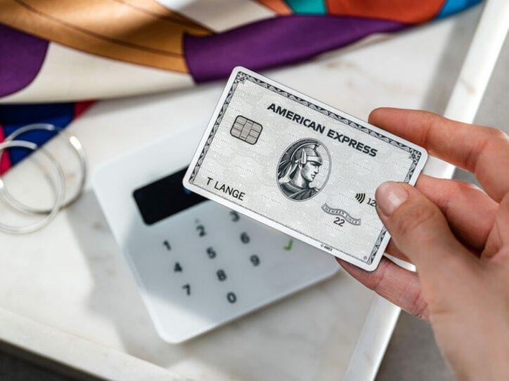 Nahaufnahme einer Hand, die eine silberne American Express Kreditkarte an ein Kartenlesegerät neben Modeaccessoires hält.